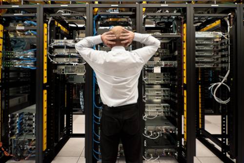 A man in white shirt and black pants standing next to server racks.